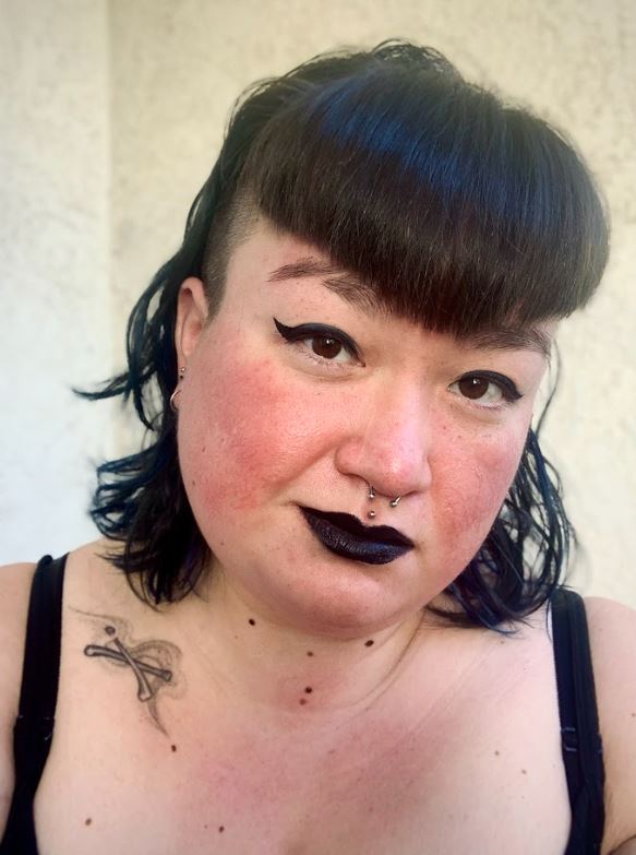 Portrait of the artist. Woman with an undercut, black bangs, black winged eyeliner, and black lipstick. She has a septum and medusa piercing and a tattoo on her collarbone.